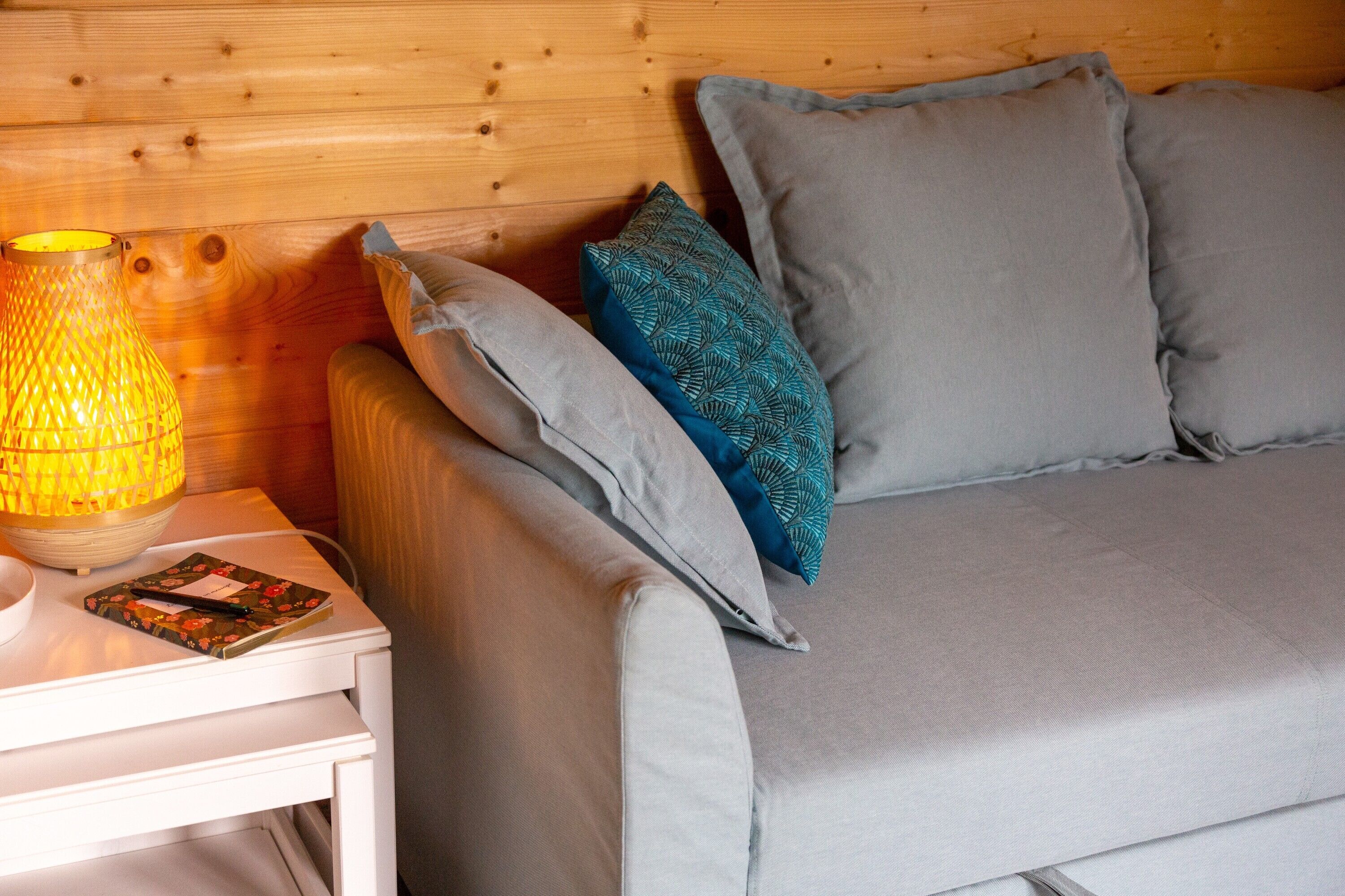 A wooden headboard, a grey sofa with pillows, a white nightstand with a lamp and a book.