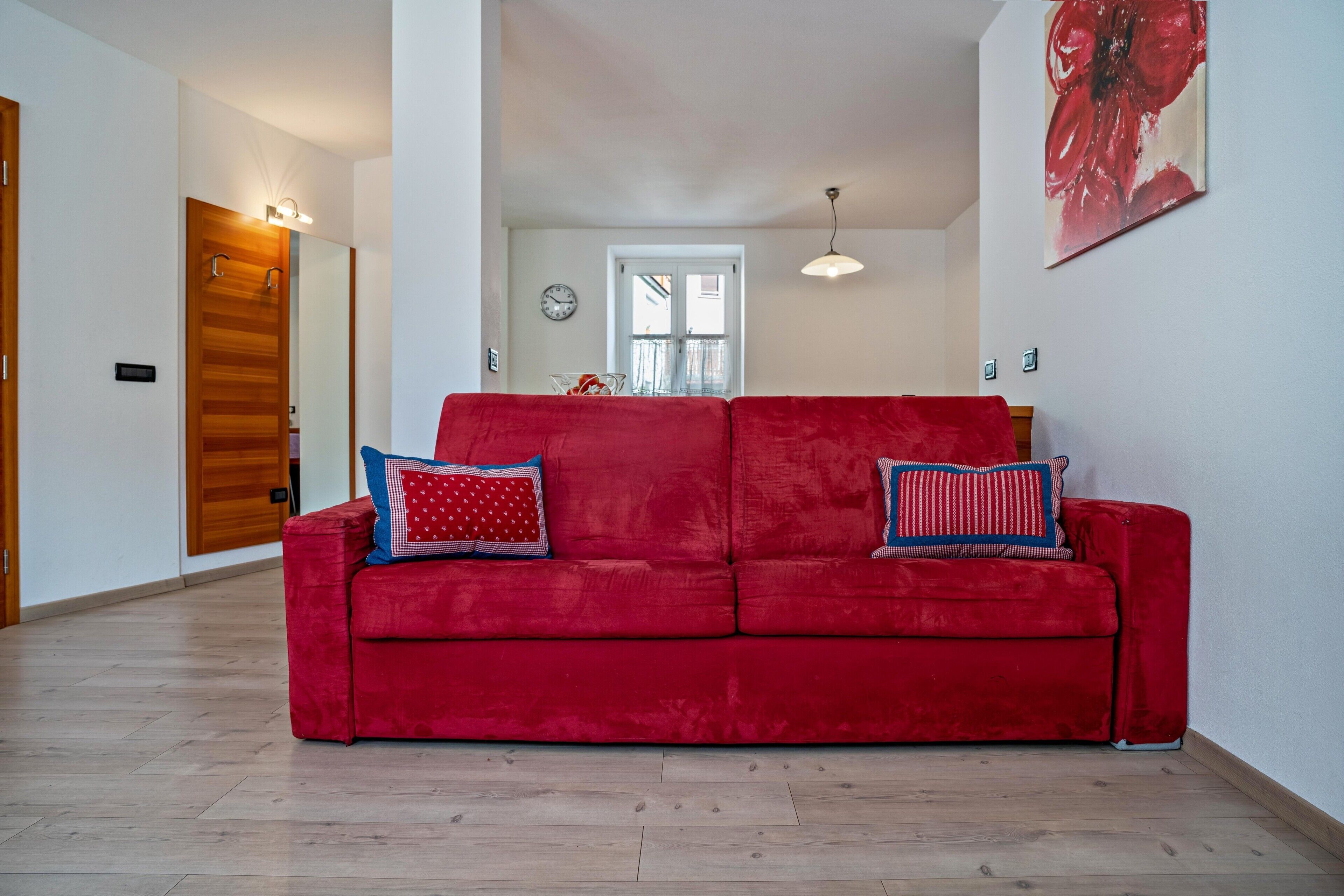 A red sofa with patterned cushions in a room with wooden flooring, a dining table with chairs, and a large window.