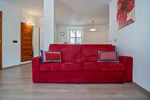 A red sofa with patterned cushions in a room with wooden flooring, a dining table with chairs, and a large window. - Apartment 'Casa Batistei Ribes' with Lake View, Balcony and Wi-Fi (Molveno)
