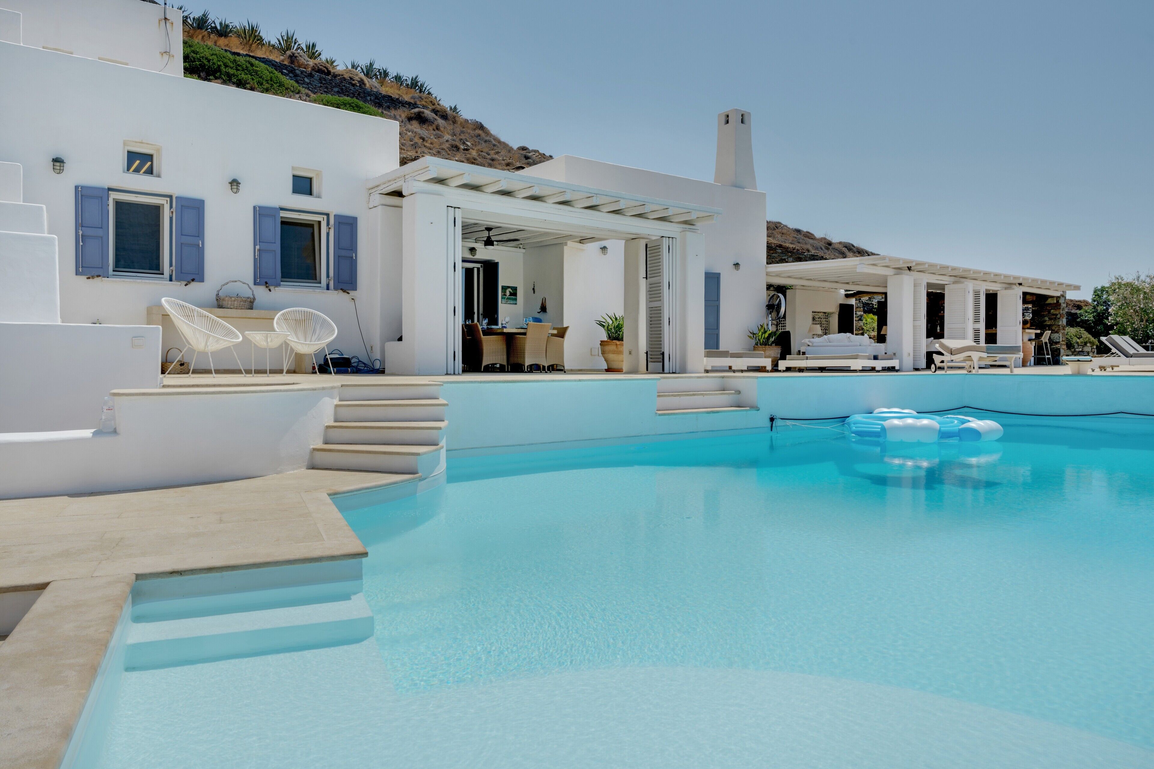 A pool area with steps leading to a covered seating area, a lounge chair, and a small table.