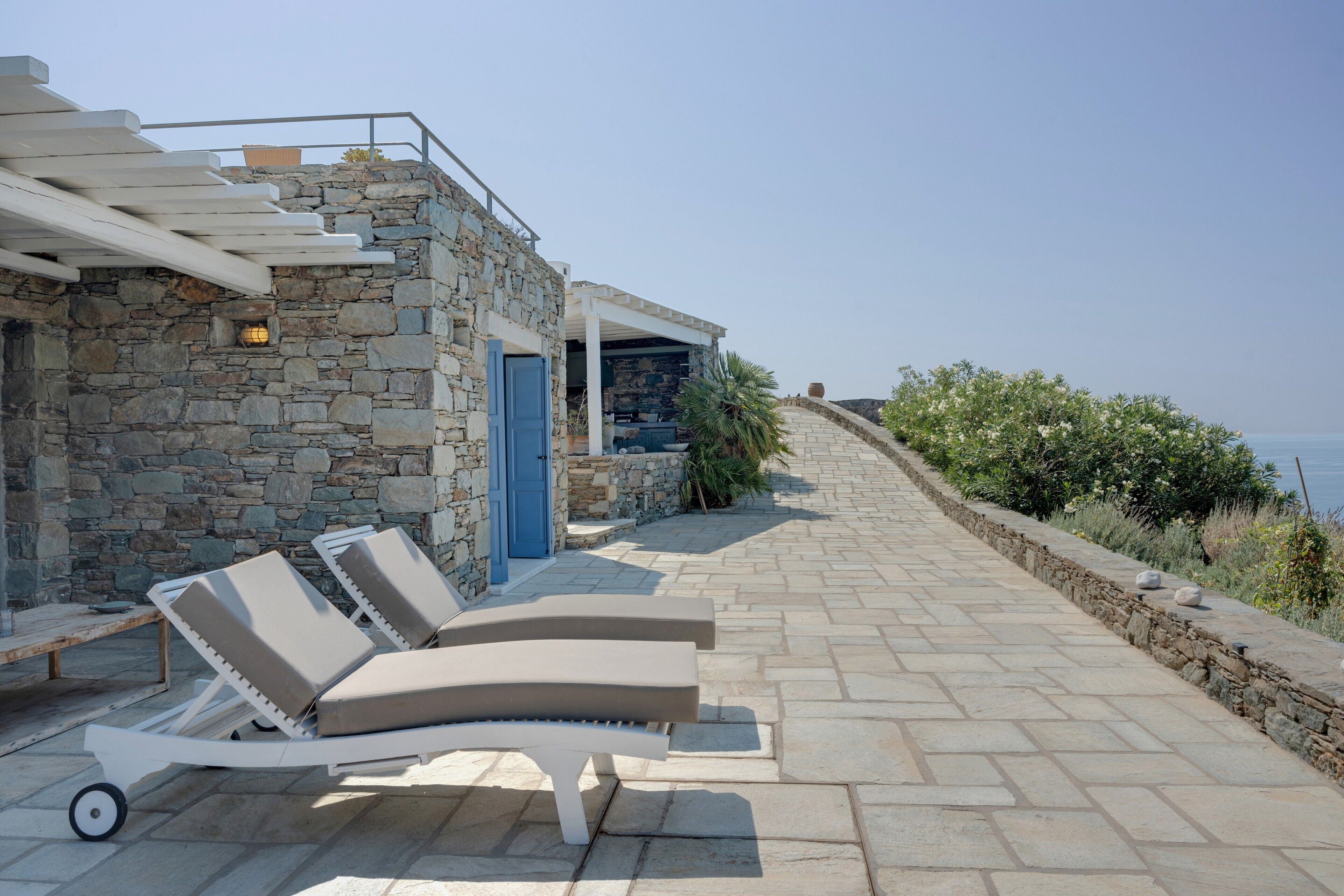 A stone building with a blue door, a covered outdoor seating area with lounge chairs, and a stone pathway leading to a scenic overlook.
