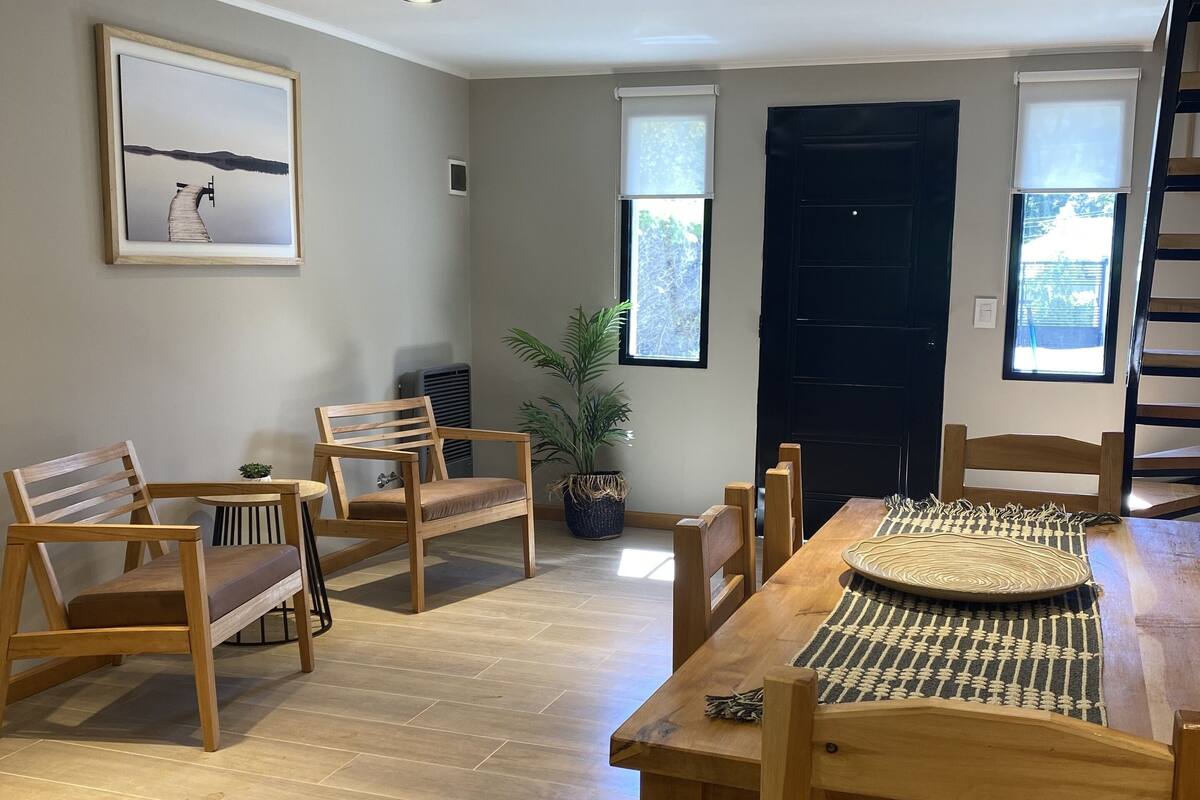 Amplia sala de estar con una mesa de comedor de madera, varias sillas de madera, un ventanal con vistas, un cuadro en la pared y luces empotradas en el techo.
