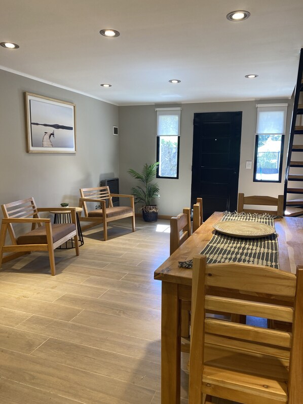 A spacious living room with a wooden dining table, several wooden chairs, a large window with a view, a framed picture on the wall, and recessed ceiling lighting. - Quillaipe Apart Suite (San Carlos de Bariloche)