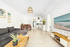 A modern living room with a sofa, coffee table, and TV. There is a dining area with chairs and a table. The kitchen is visible in the background. - Mamad Sea Breeze Netanya Retreat by SeaNRent (Netanya)