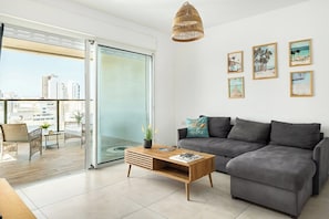A modern living room with a grey sofa, a wooden coffee table, and a city view through a glass door.