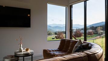 A modern living room with a large window, a leather sofa, a small round table, and a flat-screen TV mounted on the wall.