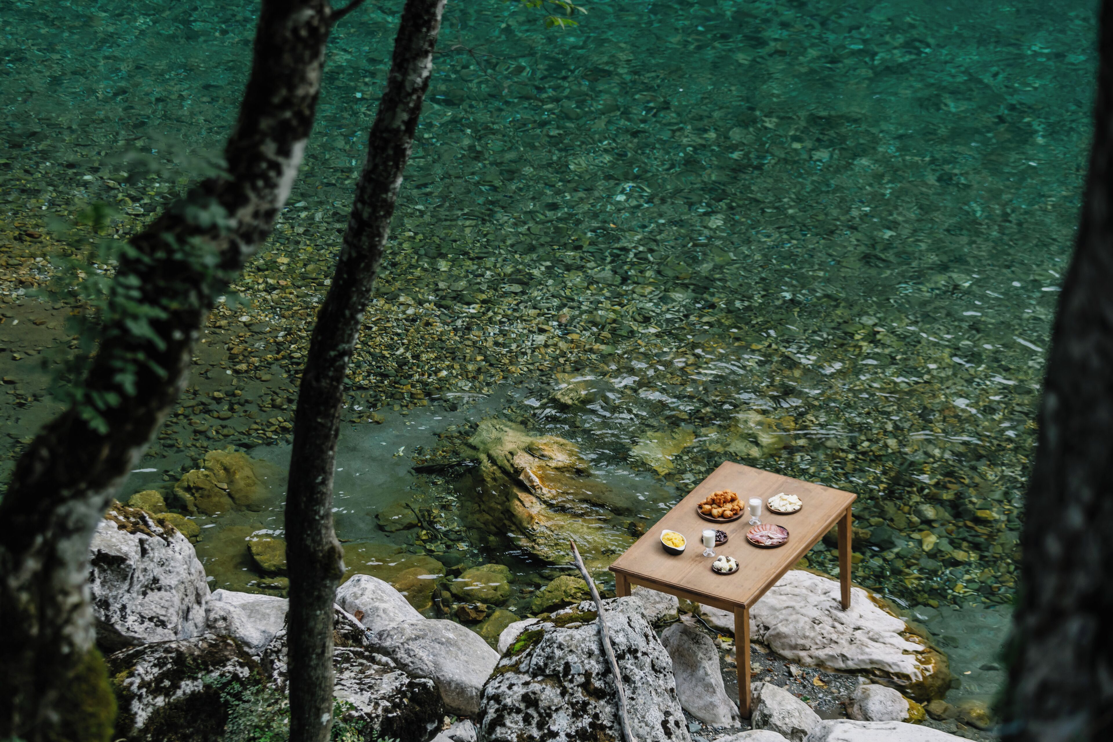Una mesa con comida sobre una roca junto a un cuerpo de agua transparente rodeado de árboles.