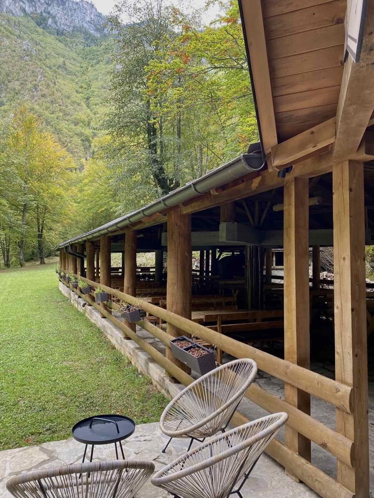 Un espace extérieur aménagé en bois, avec des tables et des chaises, entouré d’une pelouse verdoyante et d’arbres.