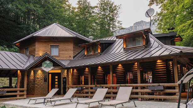 Ein Hotel in Blockhausbau mit einer überdachten Terrasse, Liegestühlen und einem Blick auf die Berge.