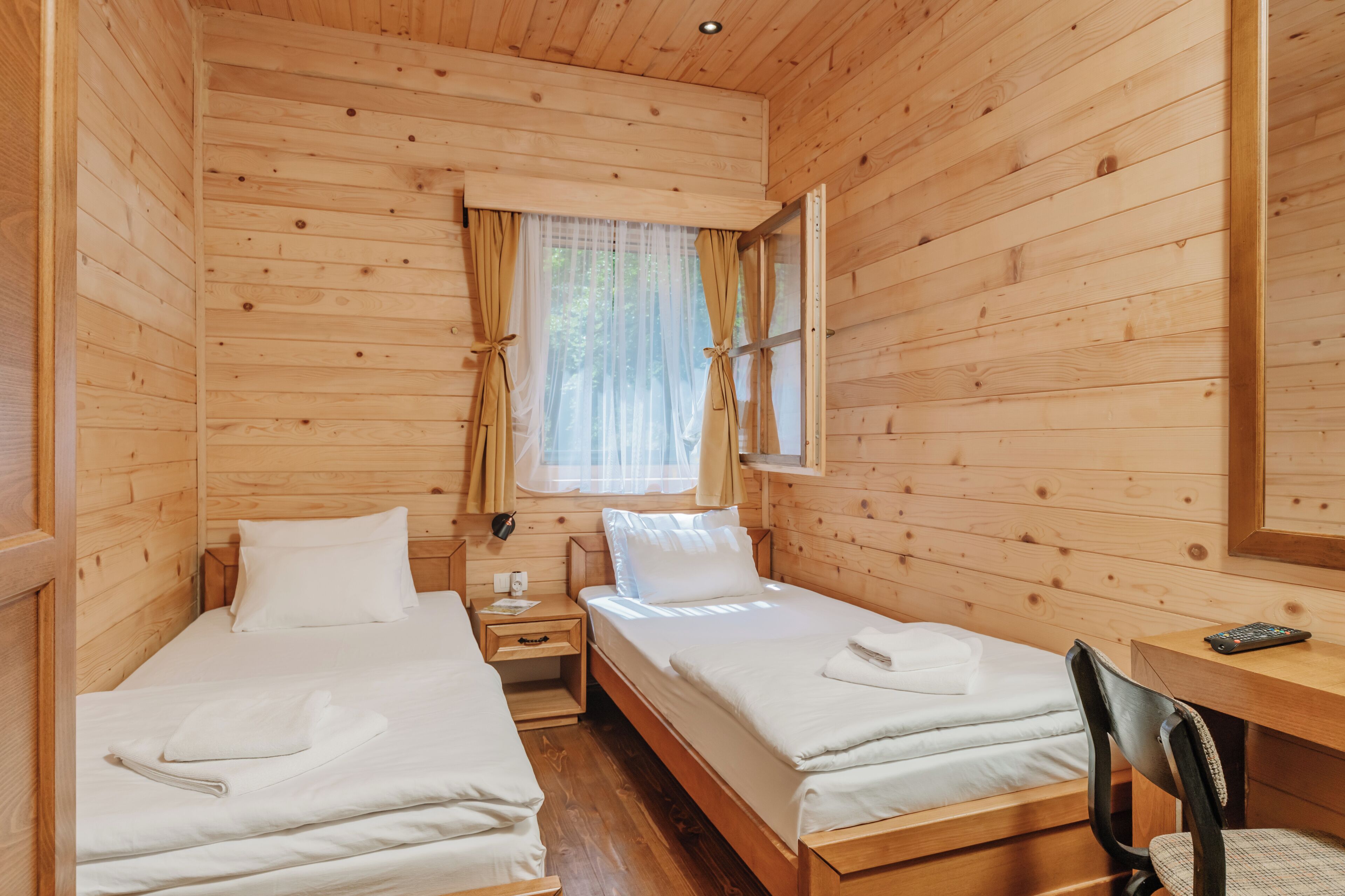 A wooden cabin room with two beds, a desk, and a window with curtains.