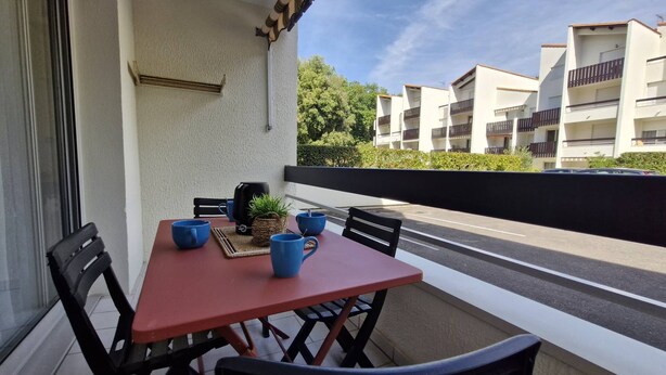 Ein Balkon mit einem roten Tisch, zwei blauen Stühlen, einem schwarzen Grill und einem kleinen Topfpflanzen.