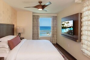 A hotel room with a bed, a ceiling fan, a TV mounted on the wall, and a window with a view of the ocean. - Waikiki | Signature Beach Escape | Pool, Dining & Sunset Walks (Honolulu Oahu)