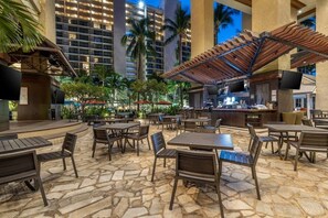 An outdoor patio area with stone flooring, wooden tables and chairs, a bar area, and palm trees. - Oahu | Oceanview Coastal Stay | Resort Pool & Beach (Honolulu Oahu)
