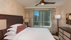 A hotel room with a bed, two bedside lamps, a ceiling fan, a large window with curtains, and a view of high-rise buildings.