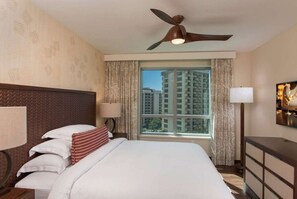 A hotel room with a bed, two bedside lamps, a ceiling fan, a large window with curtains, and a view of high-rise buildings. - Waikiki | Island Comfort Stay | Pool & Beach Access (Honolulu Oahu)