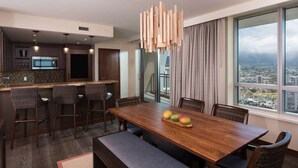 Une chambre d’hôtel moderne dotée d’un coin repas avec une table en bois et des tabourets de bar, d’une grande fenêtre offrant une vue sur la ville et d’un luminaire suspendu contemporain.