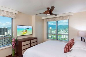 A hotel room with a bed, a TV on the wall, a ceiling fan, and large windows with a city and mountain view.