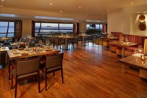 A restaurant with wooden flooring, tables set for dining, and large windows offering a view of the sea.