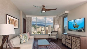 A modern hotel room with a large window offering a city view, a ceiling fan, a sofa with patterned cushions, a coffee table, and a TV on a wooden cabinet. - The Grand Islander, a Hilton Grand Vacations Club - 1 Bedroom - Mountain View - 1BR (Honolulu Oahu)