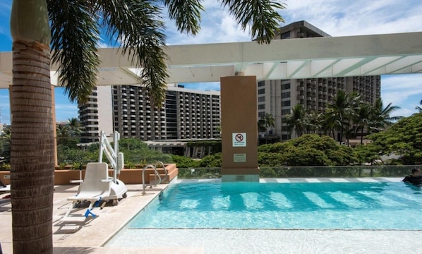 Una piscina con sillas de descanso, palmeras y un edificio al fondo.