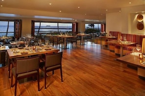 Un restaurante con suelo de madera, mesas dispuestas para comer y amplias ventanas con vistas al mar.