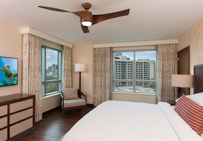 Habitación de hotel con cama, una silla, escritorio, ventilador de techo y ventanas amplias con vista a la ciudad.