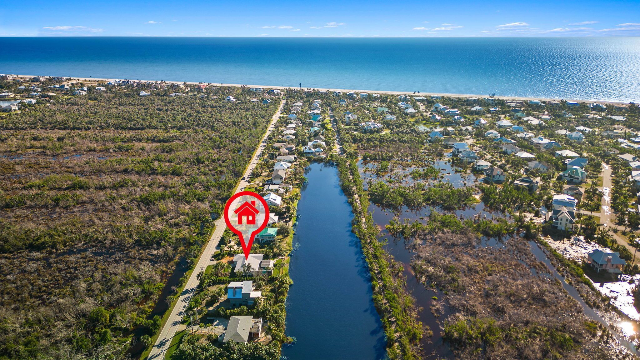 An aerial view of a coastal residential area with houses along a narrow waterway, surrounded by dense vegetation and a beach in the background.