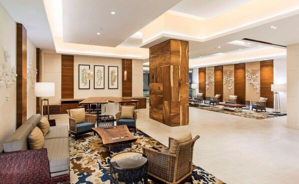 A spacious lobby with a central wooden column, seating area with sofas and chairs, and framed artwork on the walls. - Waikiki | Partial Ocean View | Pool & Beach | 2BR Plus (Honolulu Oahu)