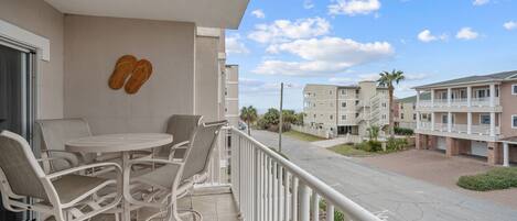 A balcony with a round table and chairs, a pair of slippers on the wall, and a view of other buildings and a palm tree.