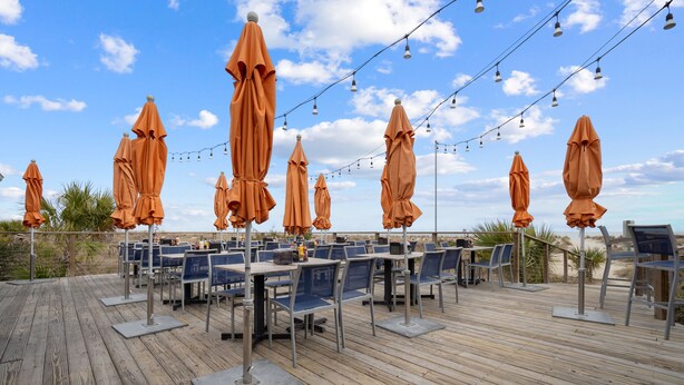 An outdoor dining area with tables and chairs, covered by orange umbrellas, and string lights overhead. - Tybee Beachfront Condo, 2 Pools & Restaurant, 1–3BR Flexibility, Turtle Time (Tybee Island)