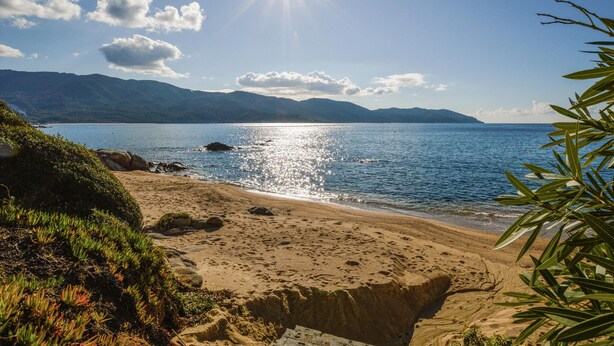 Ein Strand mit blauem Himmel, Bergen im Hintergrund und Sonnenlicht, das sich auf dem Wasser spiegelt.