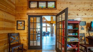 A wooden cabin interior with a large open door leading to a dining area, a bookshelf with various items, and a sign indicating a pool hall.