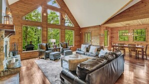 A spacious log cabin interior with a high ceiling, large windows, a fireplace, leather furniture, and a dining area.