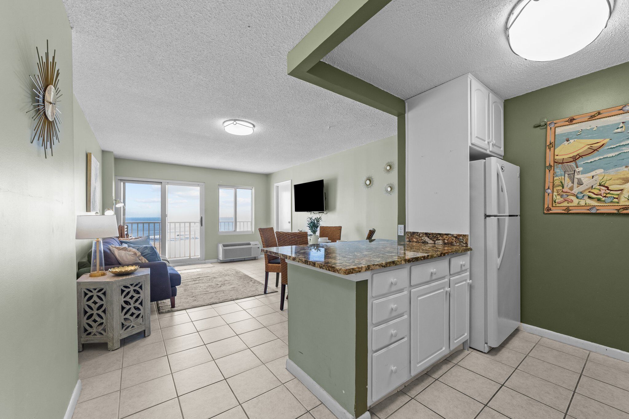 A kitchen with a large island, white cabinets, and a refrigerator. There is a dining area with a table and chairs, and a view of the ocean through a sliding glass door.