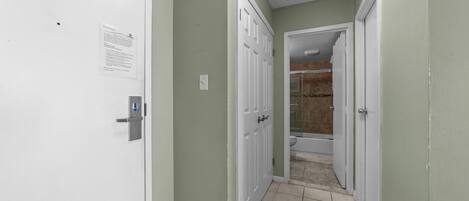 Un couloir étroit avec des portes blanches, un sol carrelé et une salle de bain visible, équipée d’une douche et de toilettes.