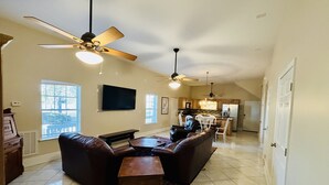 A spacious living room with a large sectional sofa, a coffee table, and a television mounted on the wall. There are ceiling fans, a kitchen area in the background, and a dining area with white chairs. - “3bdrm Barn Apt” @ Sunset Pointe (Lincoln)