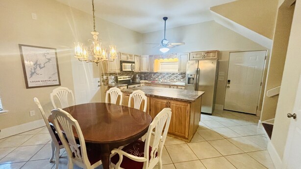A dining area with a wooden table and white chairs, a kitchen with a central island, a ceiling fan, and a refrigerator. - “3bdrm Barn Apt” @ Sunset Pointe (Lincoln)