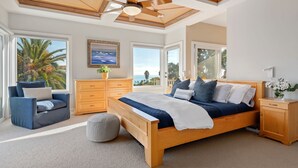 A bedroom with a large bed, wooden headboard, a ceiling fan, a nightstand, a dresser, and a view of palm trees and the ocean through large windows.