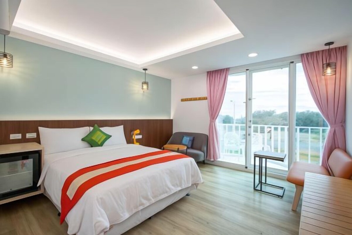 A hotel room with a large bed, a desk, a chair, and a balcony with curtains.