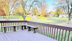 Une terrasse en bois avec des bancs, entourée d’une cour clôturée, agrémentée d’arbres et avec un bâtiment visible au loin.
