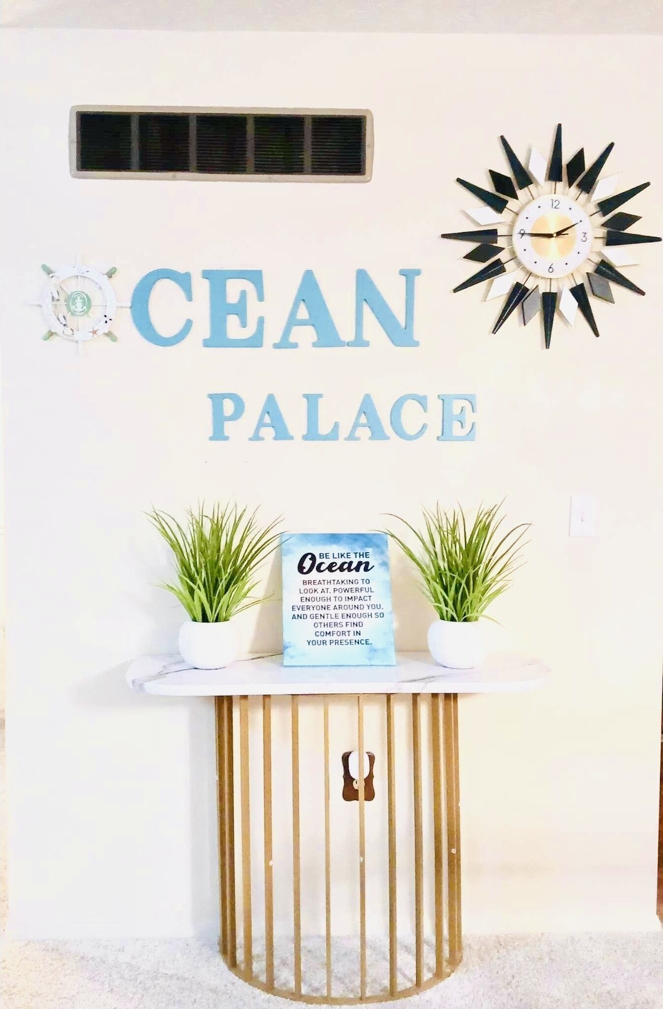 En væg med teksten "CEAN PALACE" og et dekorativt ur. Der er to plantede planter i krukker på et lille bord under teksten.