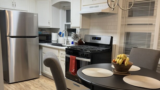 Una cocina moderna con armarios blancos, un refrigerador de acero inoxidable, una estufa negra y una mesa de comedor con sillas.