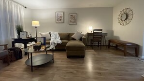 Una sala de estar con un sofá, una mesa de centro, un escritorio con una silla y un banco. Hay dos cuadros en la pared, una lámpara en el escritorio y una planta sobre la mesa de centro.