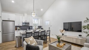 A modern kitchen with white cabinets, a stainless steel refrigerator, and a microwave. There is a dining area with a table and chairs, and a living room with a sofa and a coffee table. - Nestled Nook Bungalow #7033A (Houston)
