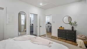 A bedroom with a large bed, a dresser, a mirror, and a view into the kitchen. - Nestled Nook Bungalow #7033A (Houston)