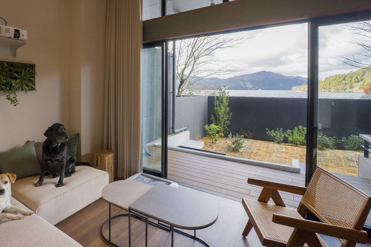 A modern living room with a sofa, a table, and a view of a garden and mountains through large windows.