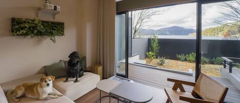 A modern living room with a sofa, a table, and a view of a garden and mountains through large windows.