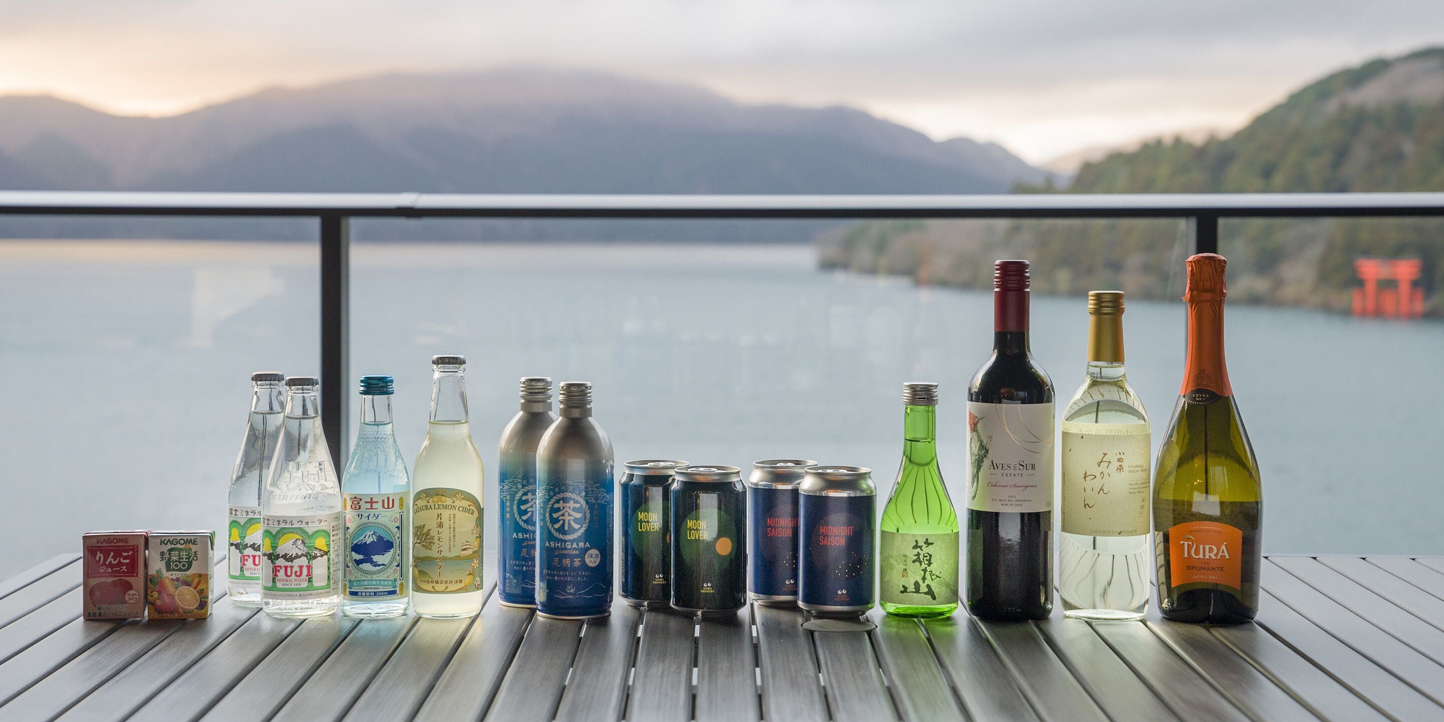 A variety of beverages on a wooden table with a scenic lake and mountains in the background.