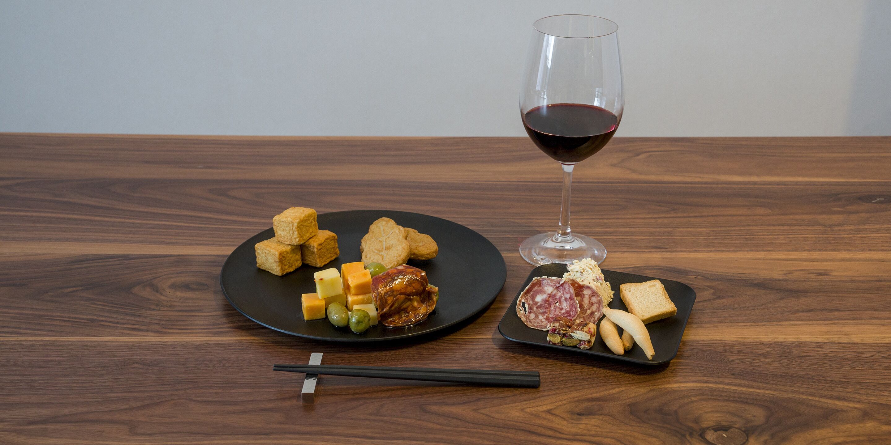 A wooden table with a black plate of food, a glass of red wine, and chopsticks.