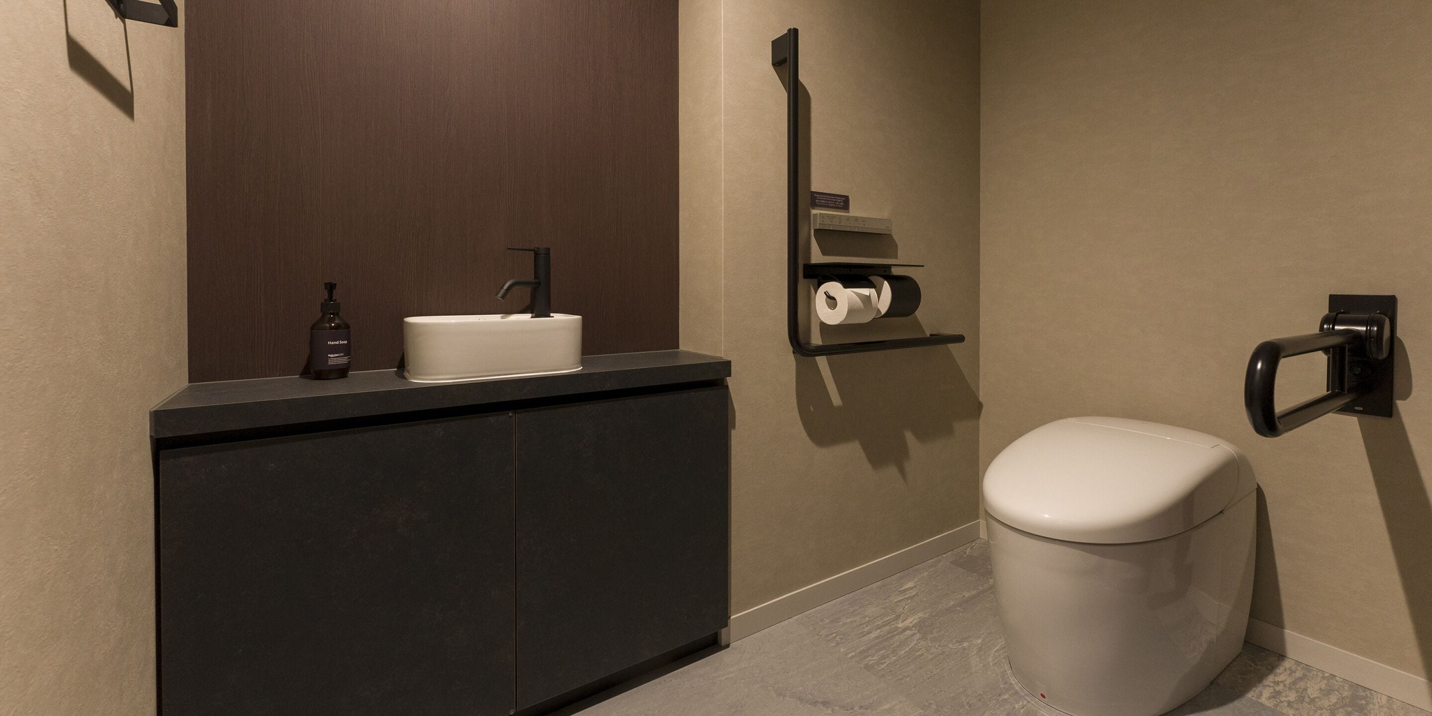 A modern bathroom with a dark countertop, a white sink, a toilet, and a towel dispenser.